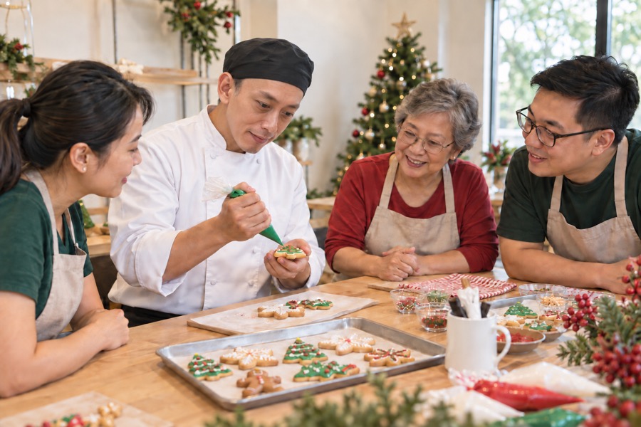 A chef piping icing onto a Christmas tree cookie while a family looks on at a decorated table of iced cookies
